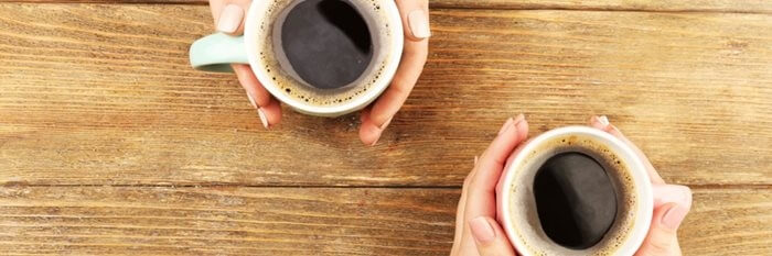 Close up of two pairs of hands, sat opposite each other, holding cups of coffee on table.
